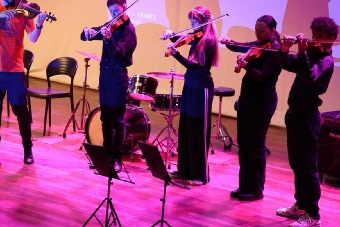 Grupo de alunos de violino em apresentação no teatro, com o público de frente
