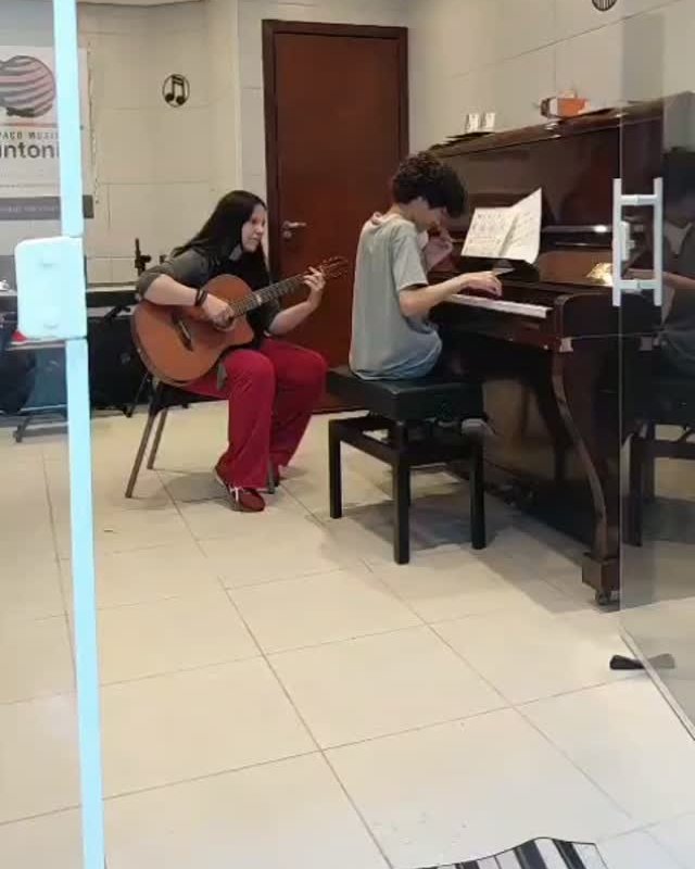 Sala da escola com aluna ao violão e aluno ao piano